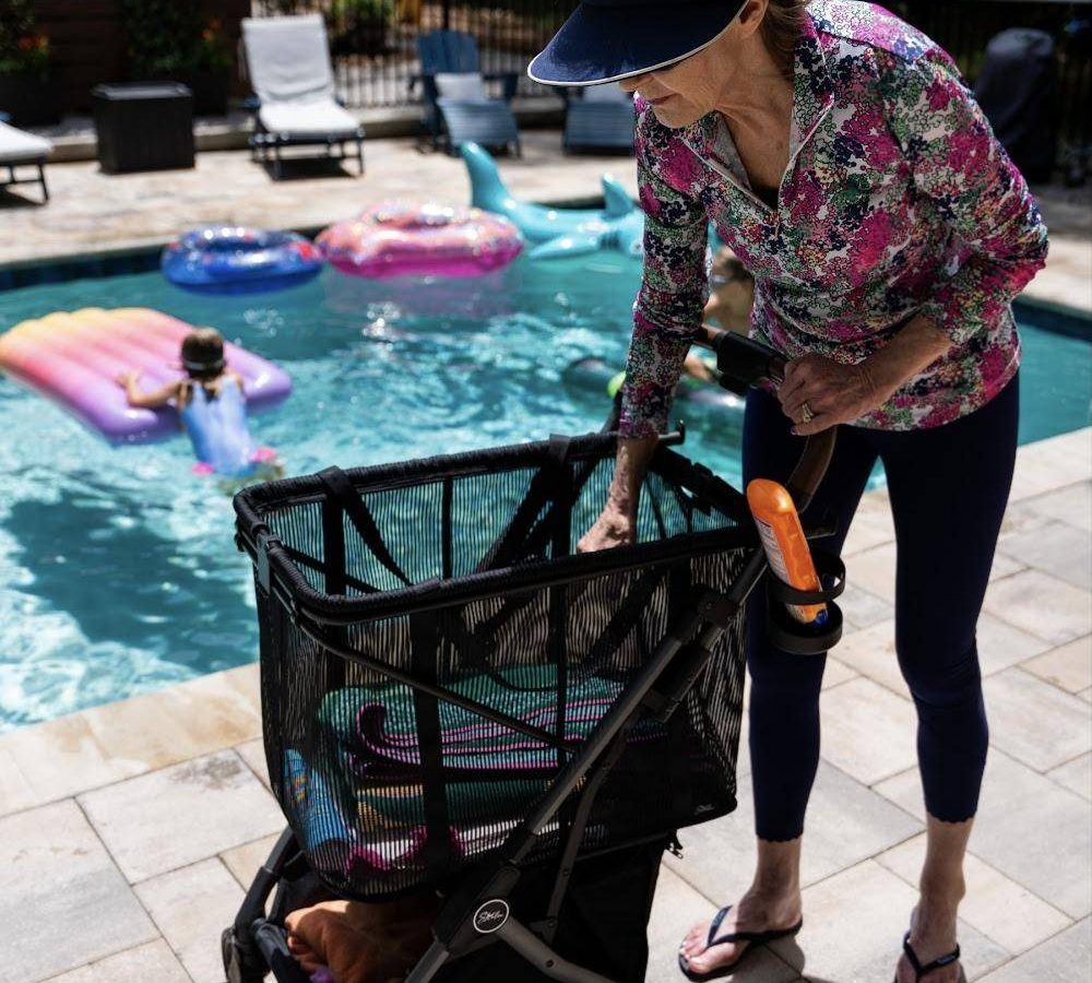 A woman beside a pool loads towels into the mesh bag of her Strolee cart
